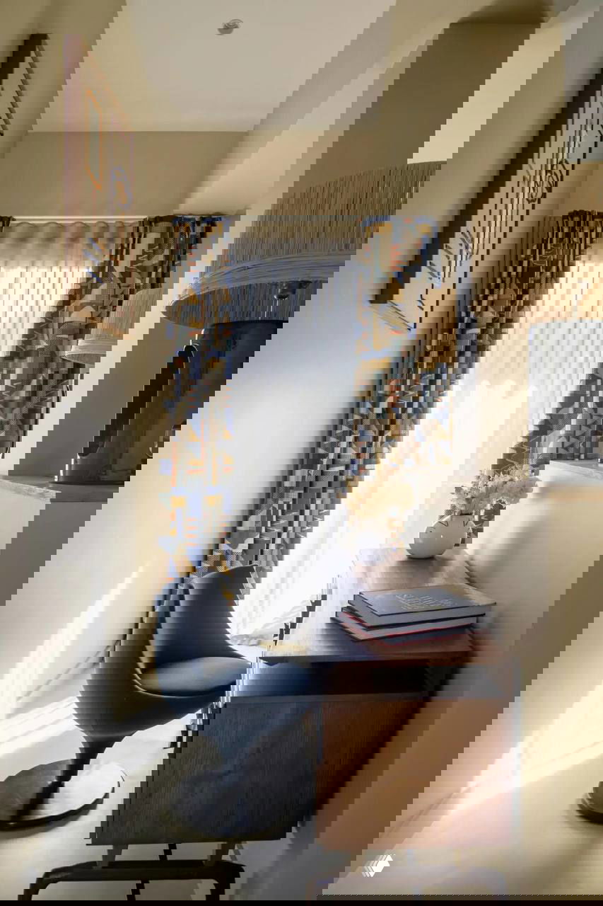 Небольшой рабочий уголок у окна: деревянный стол, современное кресло на подставке и геометрические шторы, заливаемые дневным светом.