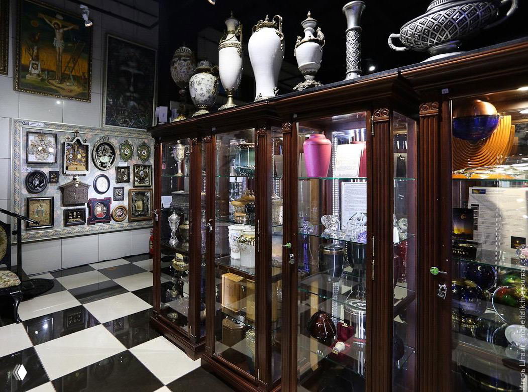 Стеклянные шкафы с урнами, вазами и мемориальными предметами на фоне чёрно-белой плитки пола в экспозиции Музея мировой погребальной культуры.