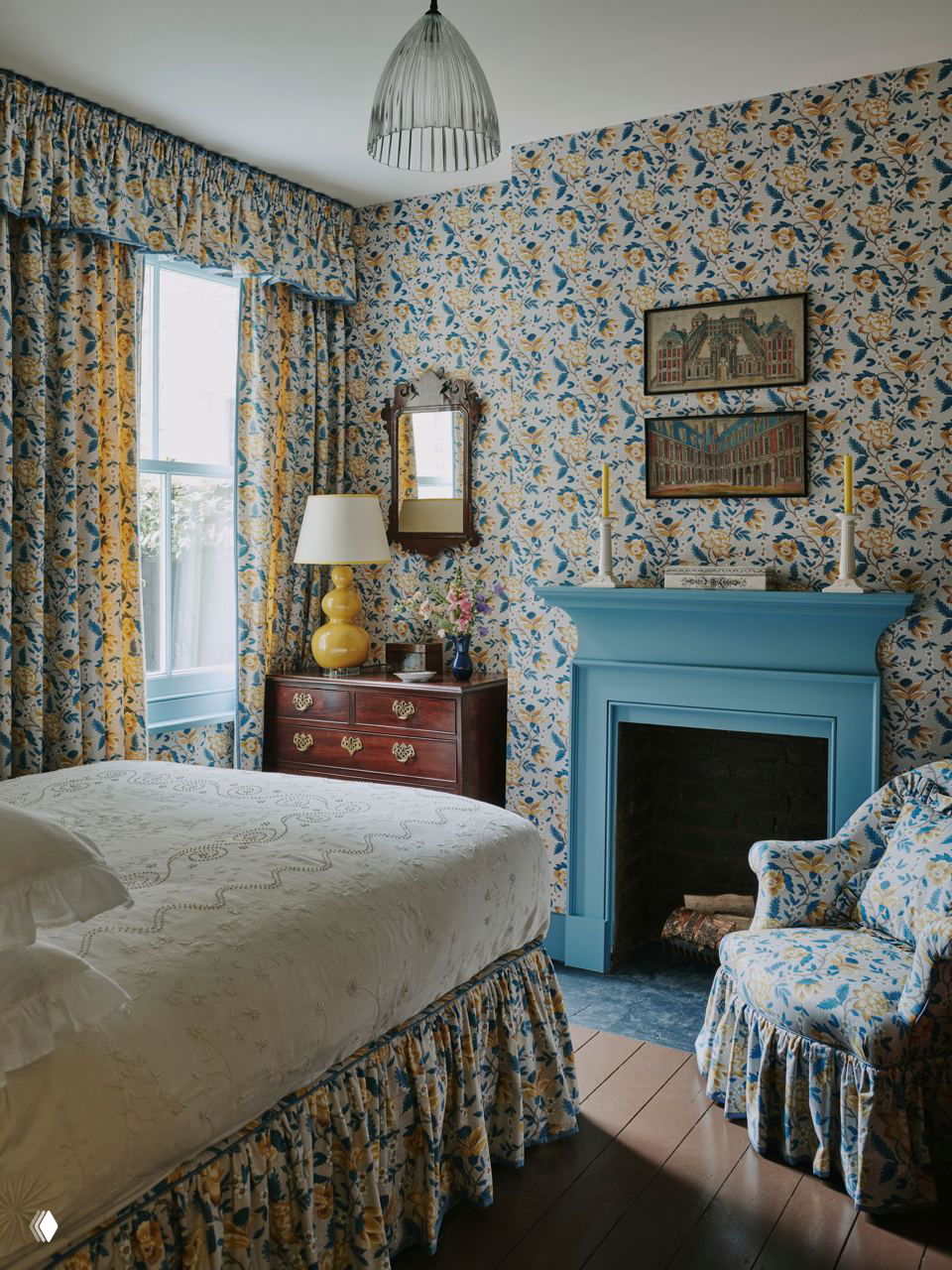 Спальня с цветочным обоями и шторами, голубым камином и классической мебелью: кровать с оборкой, комод и кресло в однотонно-цветочном оформлении.