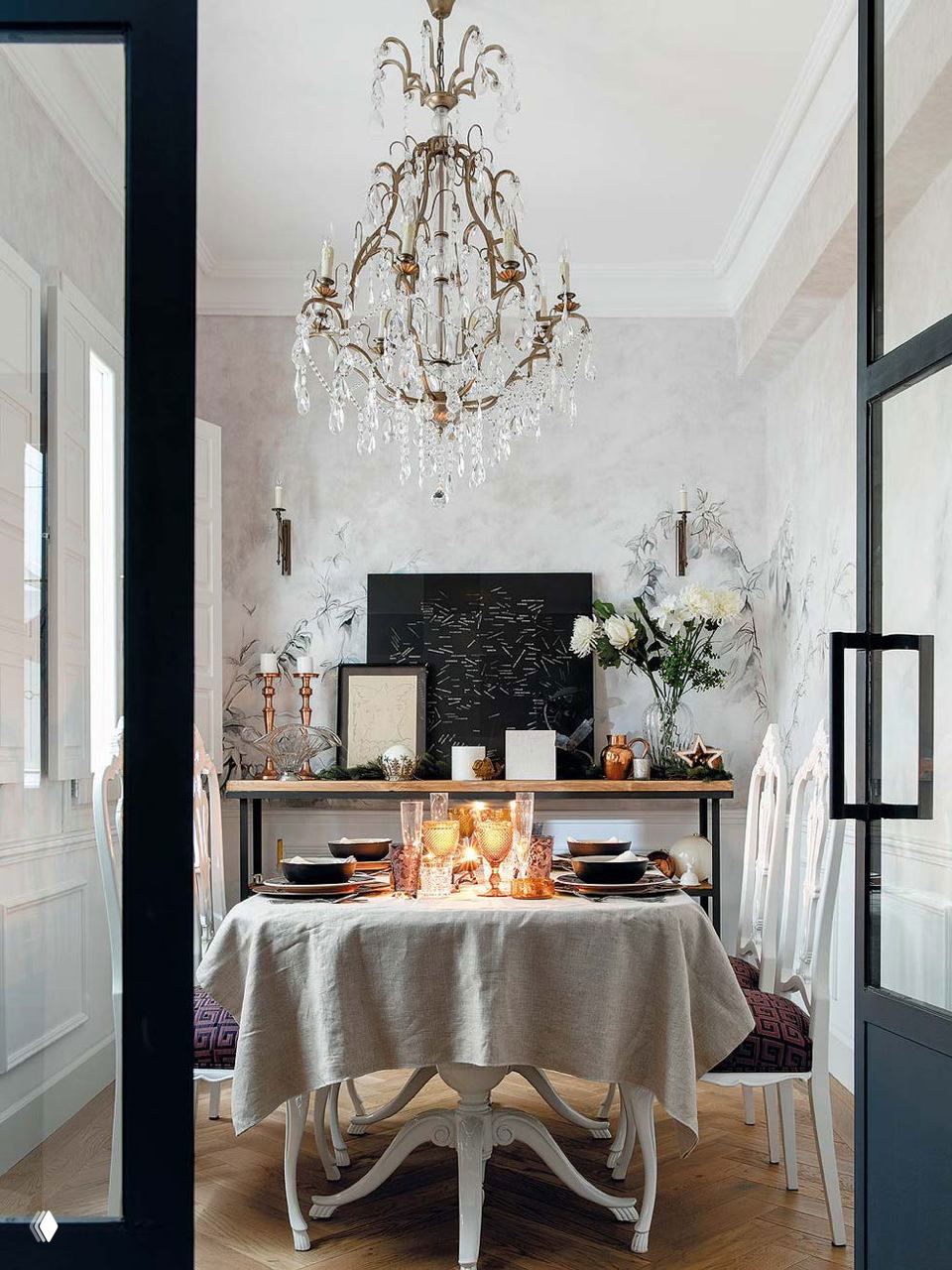 Столовая с хрустальной люстрой и сервированным столом: белые стулья, текстильная скатерть и золотистые детали на консоли за столом.