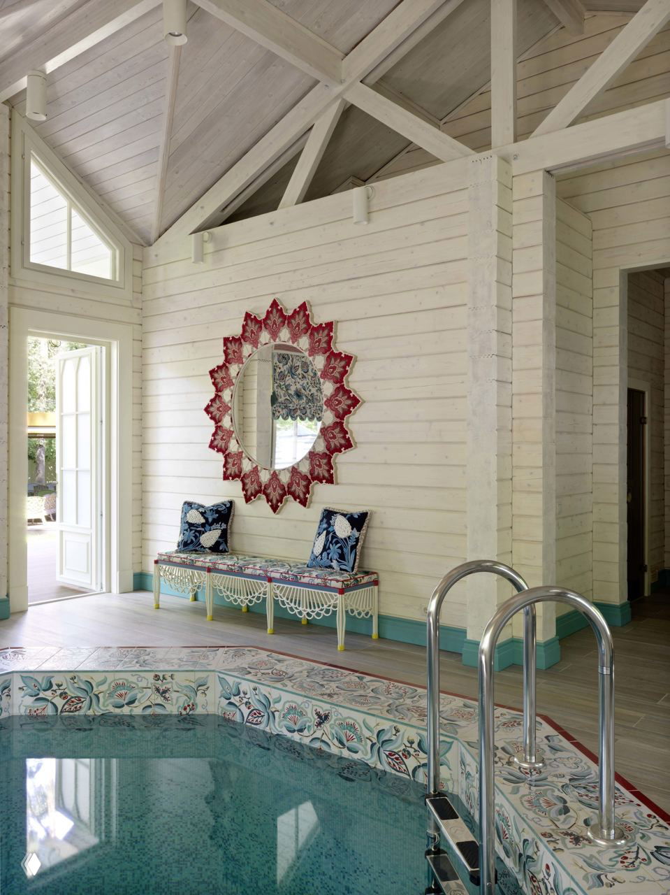 Банный зал с украшенным мозаичным бассейном, белыми окрашенными деревянными стенами, декоративным зеркалом и зоной отдыха у воды.