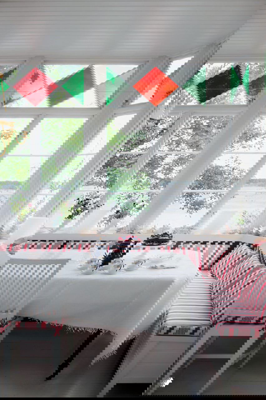 Светлая веранда-столовая с большими окнами и витражными вставками, обеденный стол со скатертью и красно-белой банкеткой у окна с видом на озеро.