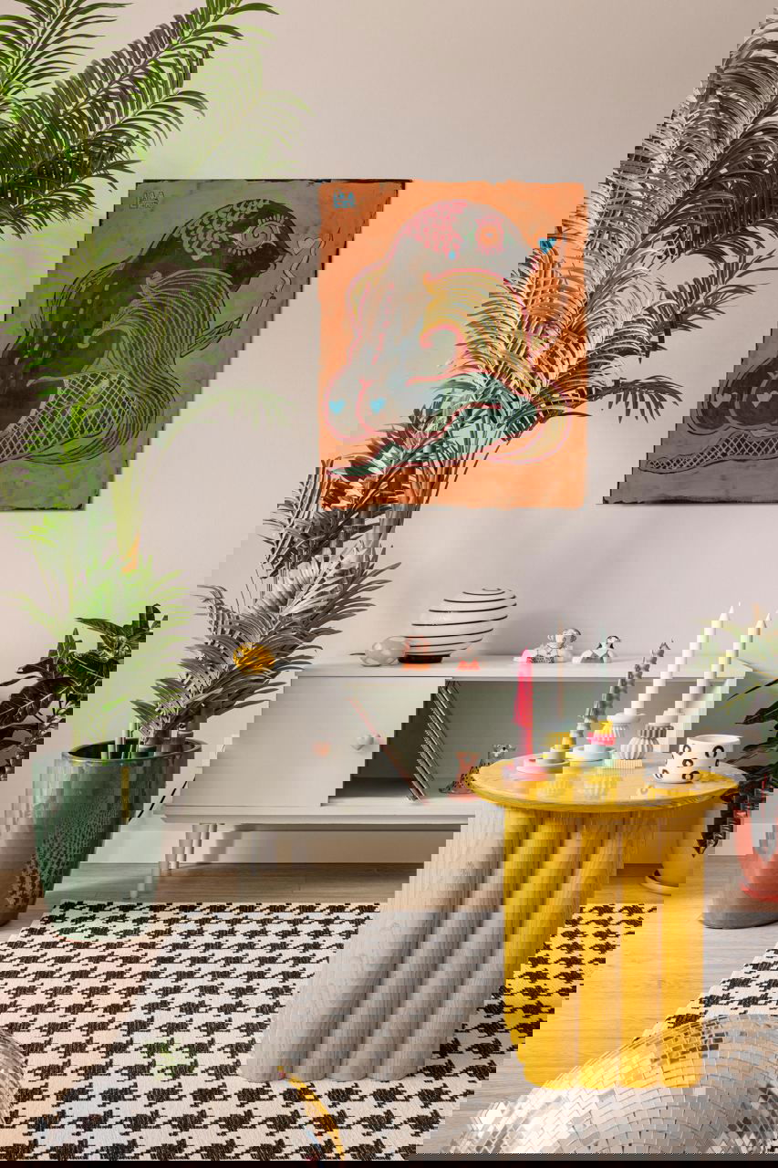 Детали гостиной: светло‑мятная тумба, настенная картина в тёплых тонах, жёлтый резной столик с набором цветных свечей и крупная зелёная пальма у стены.