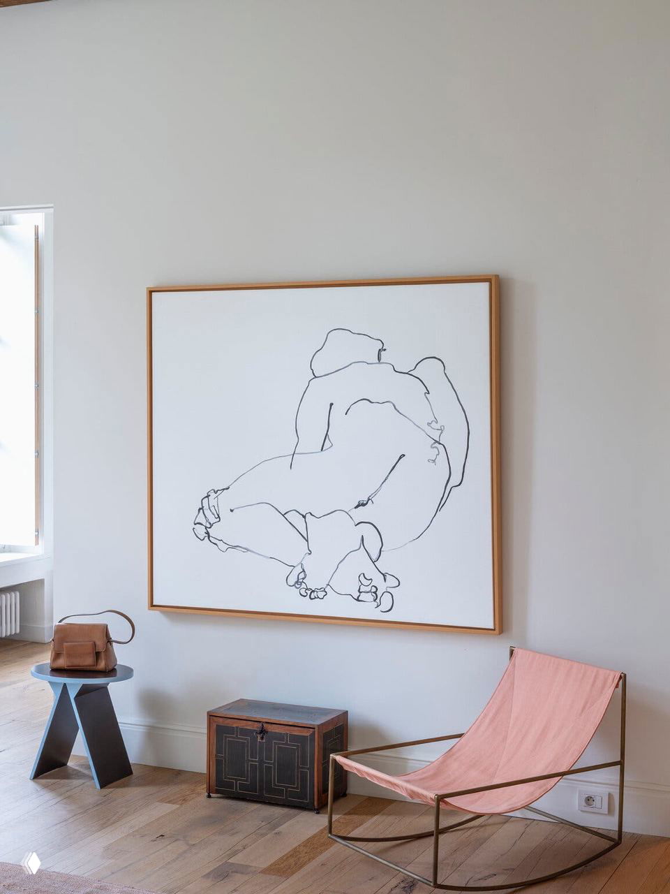 Уютный уголок с большим графическим холстом в тонкой деревянной раме, низким столиком, стулом и розовым креслом‑кресалкой на фоне деревянного пола.