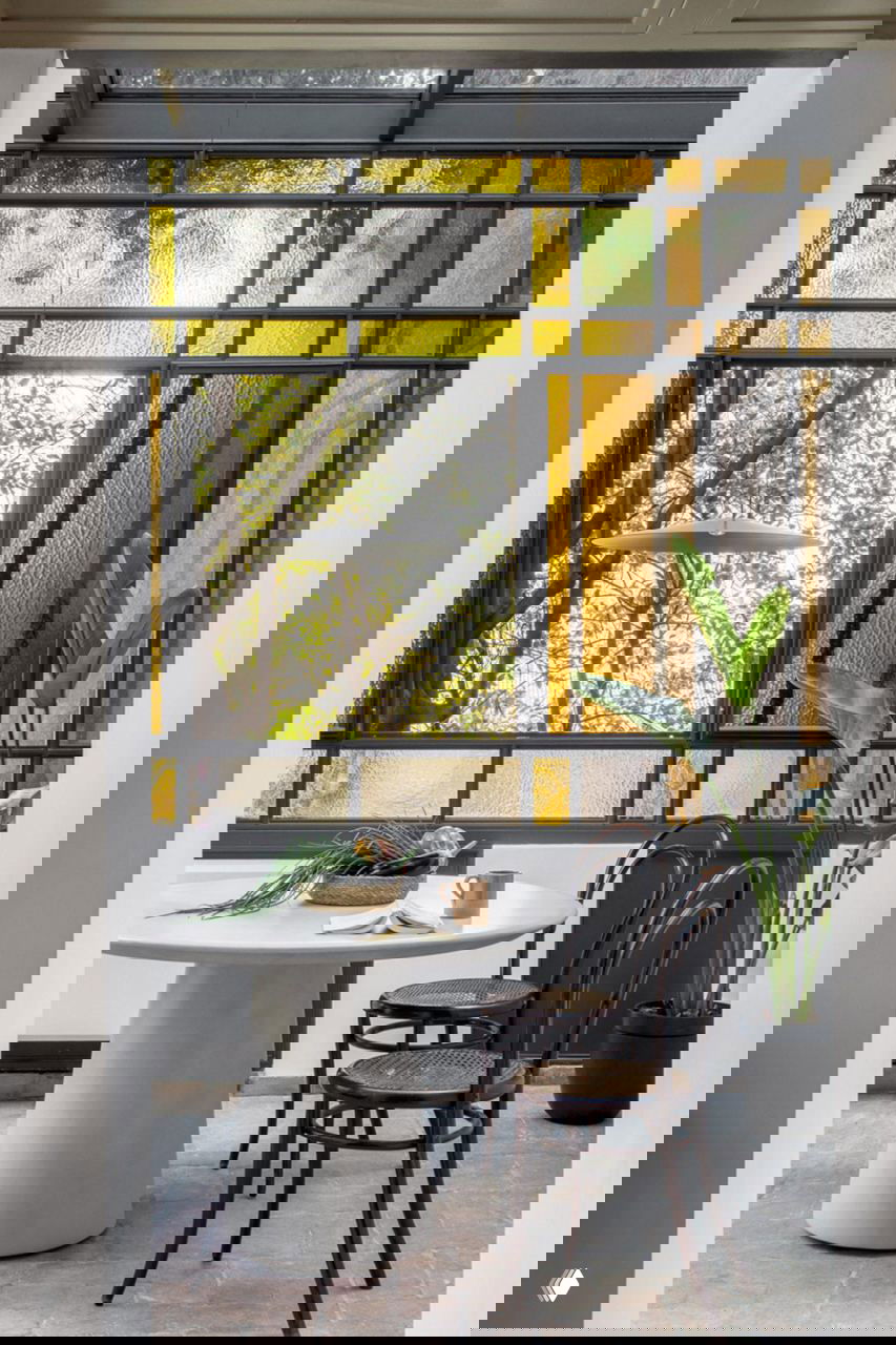 Витражный фасад с цветными стёклами формирует мягкое жёлто‑янтарное освещение в обеденной зоне рядом с круглыми столом и стульями.