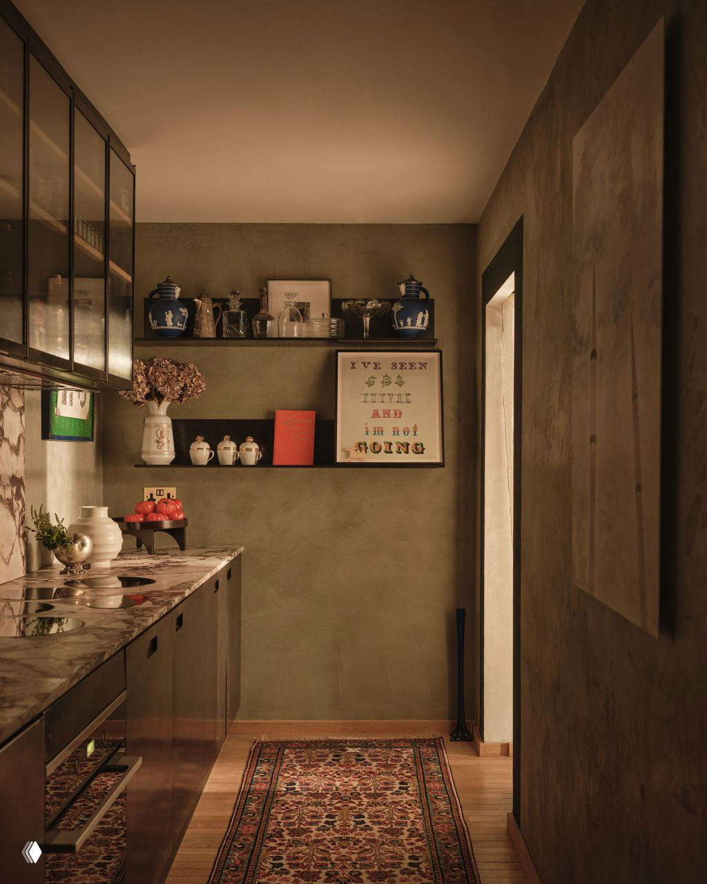 Кухонная зона с тёмными фасадами, мраморной столешницей, открытыми полками с керамикой и длинным узким ковриком на деревянном полу.