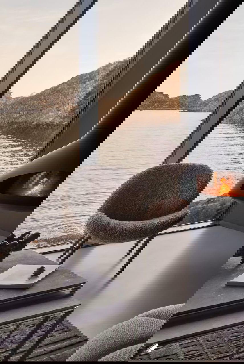 Уютный интерьер с мягким креслом у панорамного окна и подвесным камином, с видом на море и скалистые острова Лофотен.