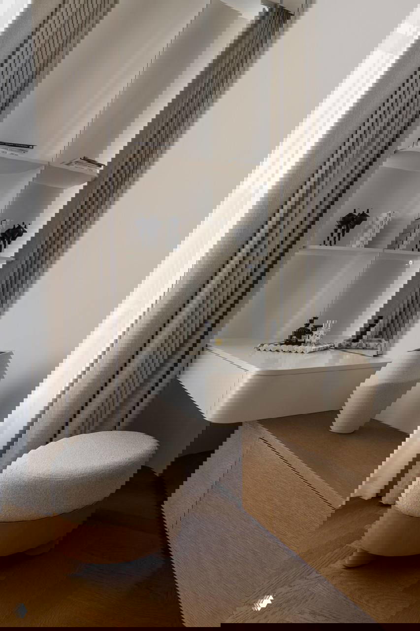 Туалетный столик и мягкий пуф у окна: минималистичный гримерный уголок с зеркалом, шторами и деревянным низким комодом в бежевой гамме.