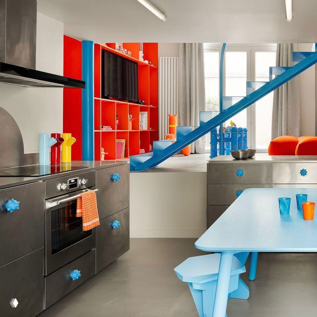 Яркая кухня-гостиная: красные открытые полки, голубая лестница, стальные шкафы и приборы, светлый голубой обеденный стол и цветные аксессуары.
