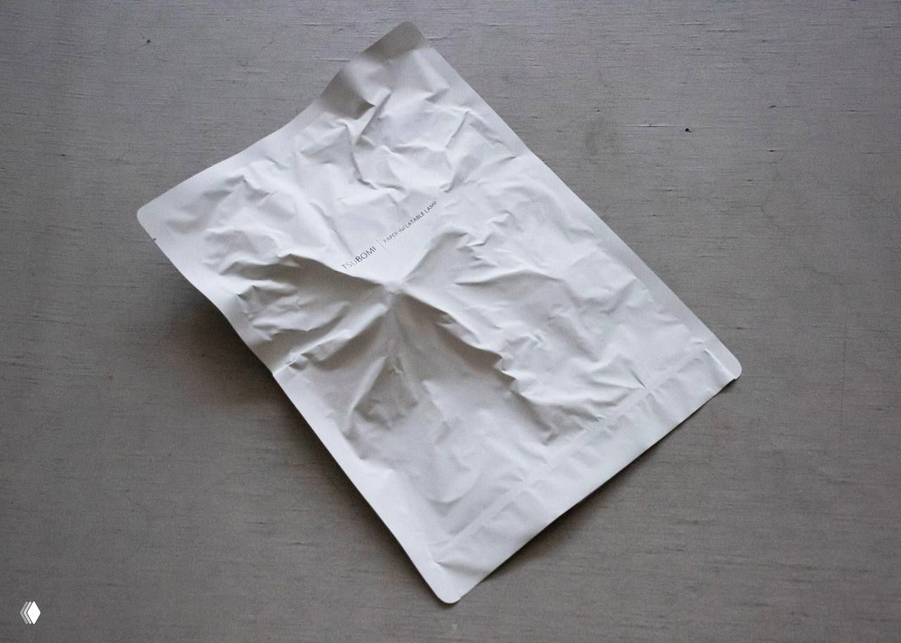 Плоская лампа Tsubomi в сложенном виде помещена в белый конверт формата A4; видно смятый бумажный пакет и компактность изделия для хранения и пересылки.