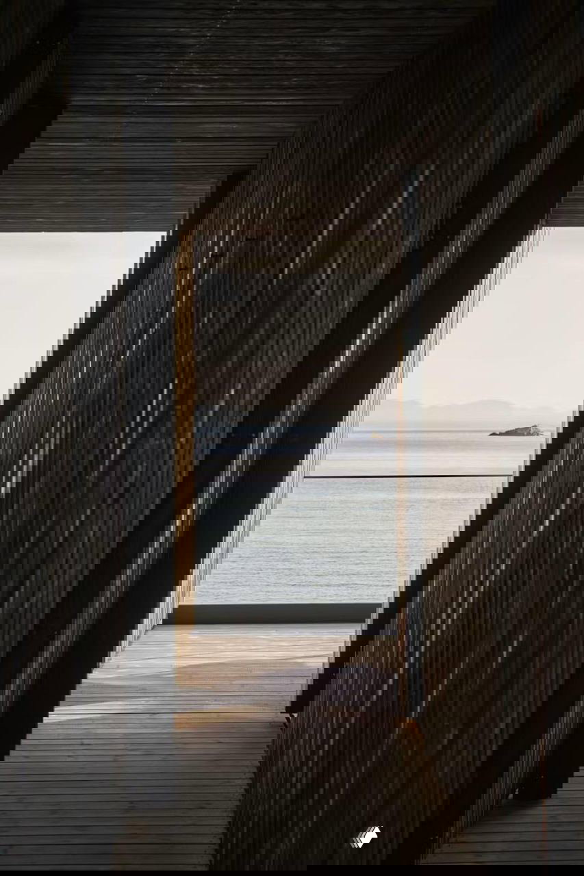 Длинный деревянный коридор между тёмными фасадами хижин, открывающий вид на спокойную морскую поверхность и дальние острова.