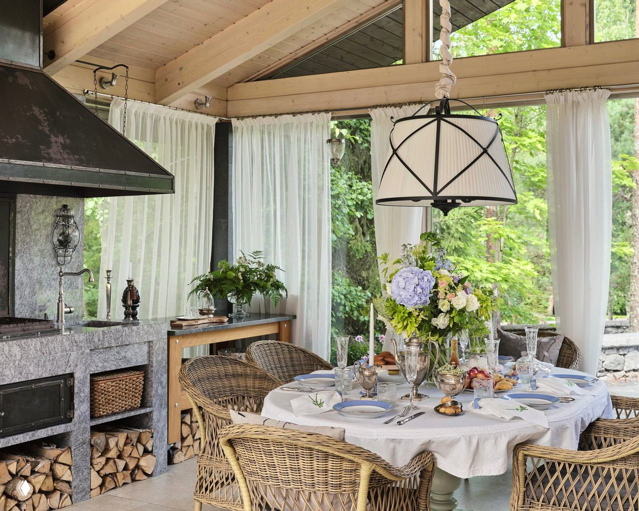 Круглый стол на веранде с плетёными стульями, белой скатертью, цветочной композицией в центре и хрустальной посудой на летней сервировке.