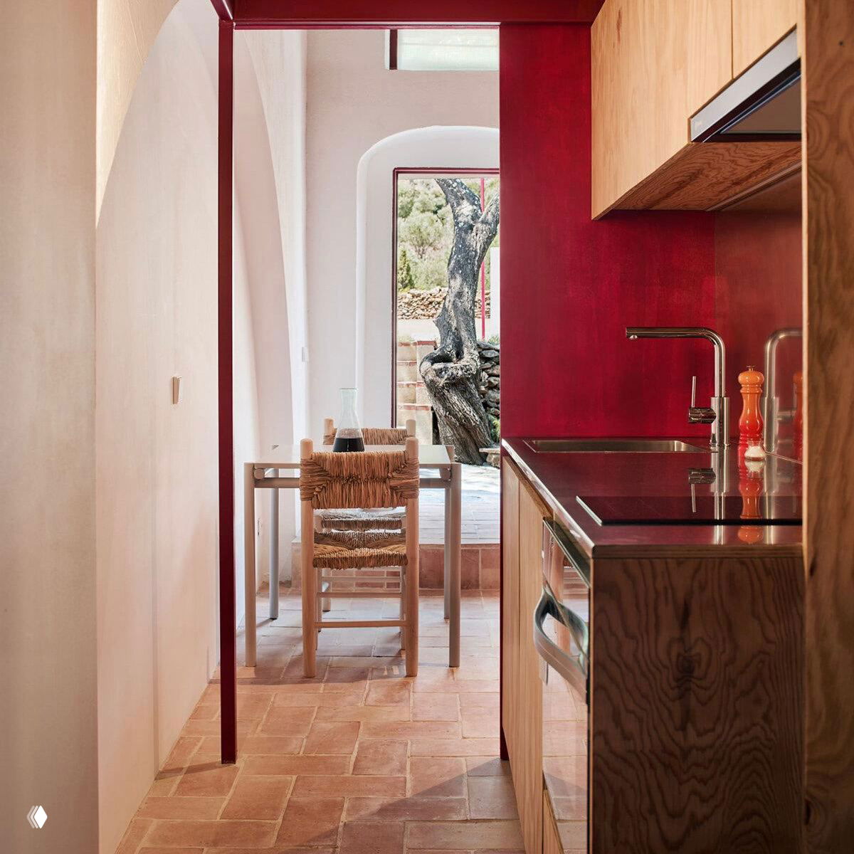 Узкая кухонная ниша с деревянными фасадами, насыщенно-красной акцентной стеной, арочным проходом, столом у окна и терракотовой плиткой на полу.