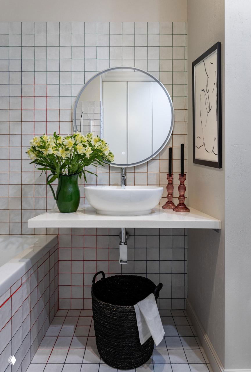 Санузел с небольшой столешницей и круглым зеркалом: плитка в сетку с цветными швами, чёрная корзина для белья и цветы в вазе для домашнего уюта.