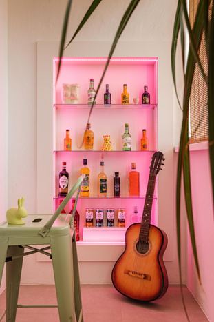Небольшой уголок с баром: встроенная полка с розовой подсветкой и бутылками, гитара в углу и декоративные предметы на фоне светлой стены, добавляющие характер пространству.