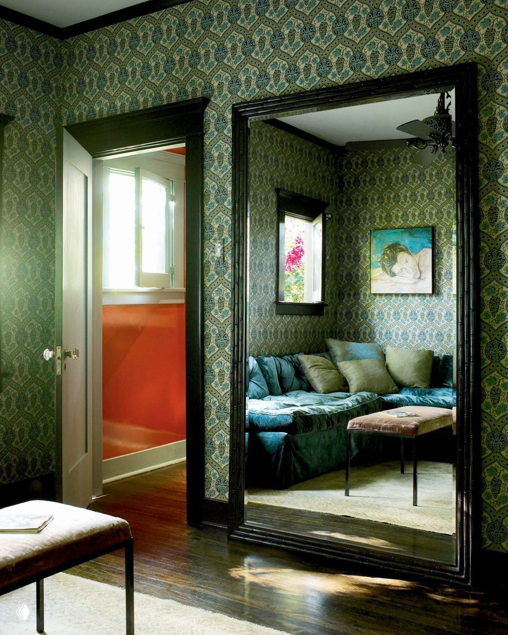 Комната с глубоким зелёным диваном на фоне узорных обоев и большим настенным зеркалом, создающим эффект увеличенного пространства и атмосферу ретро-интерьера.