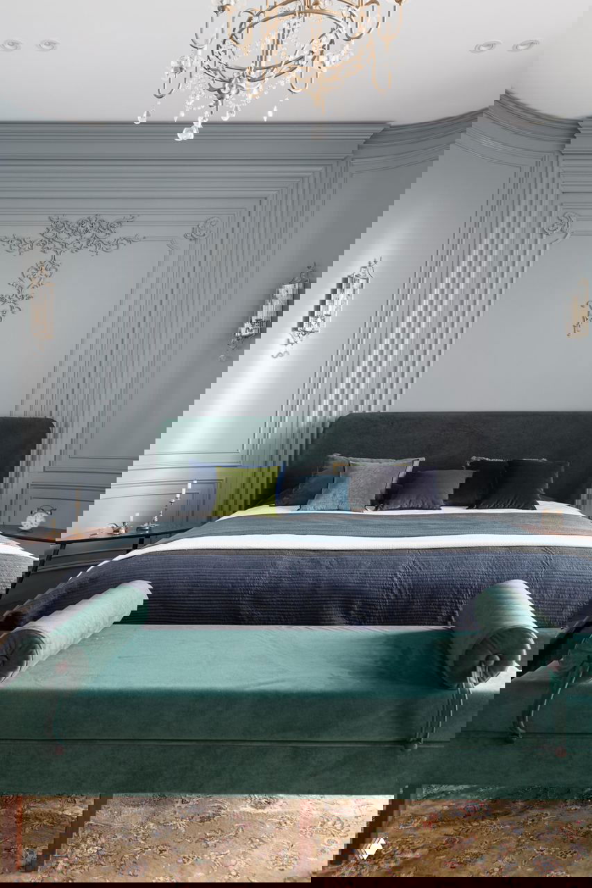 Спальня с лепниной и молдингами на стенах, велюровая изголовье тёмно-зелёного цвета и банкетка в тон — элегантный классический интерьер.