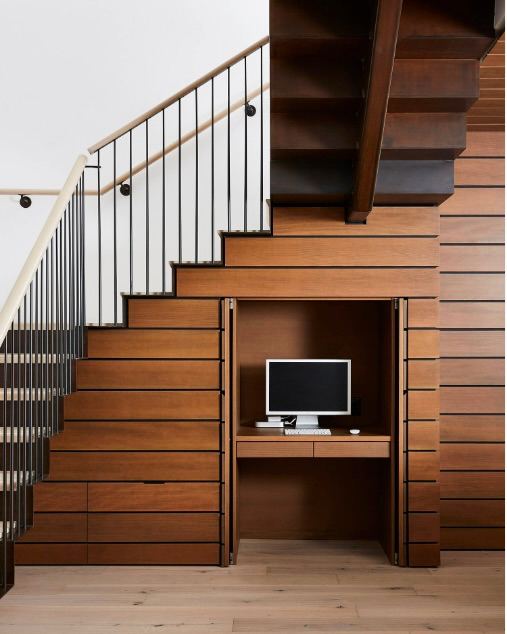 Скрытый кабинет за деревянными панелями под лестницей: небольшой стол и место для компьютера, оформление стен в тёплой древесной отделке.