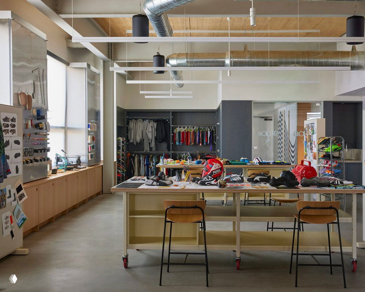 Дизайн‑мастерская Puma: большие рабочие столы, образцы обуви, инструменты и организованные стойки с материалами и макетами продуктов на фоне светлого потолка.