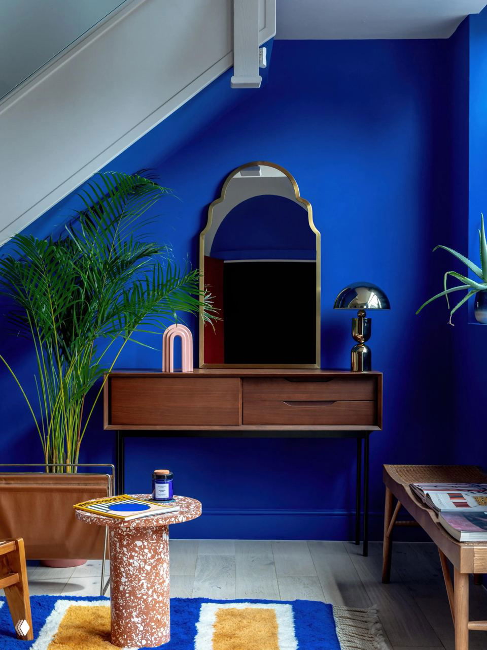 Интерьер с насыщенной кобальтово‑синей стеной, деревянной консолью, декоративным зеркалом, зелёным растением и ярким ковром с жёлтыми вставками.