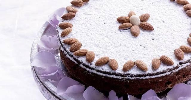 Пряный пирог, посыпанный сахарной пудрой и украшенный миндалём по краю, подан на стеклянной подставке.