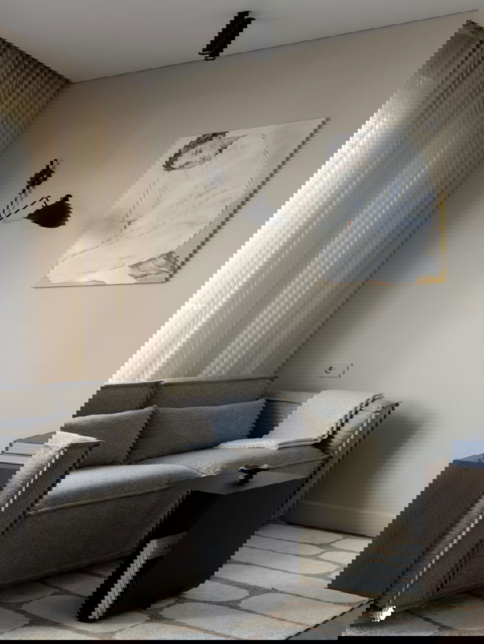 Уютная зона гостиной в проекте на 42,5 м²: серый диван, бежевые шторы, настенный светильник и маленький чёрный приставной столик на ковре.