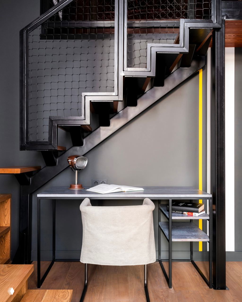 Чёрная металлическая лестница с геометричным профилем над лаконичным столом, строгая серая стена и настольная лампа создают индустриальный рабочий уголок.