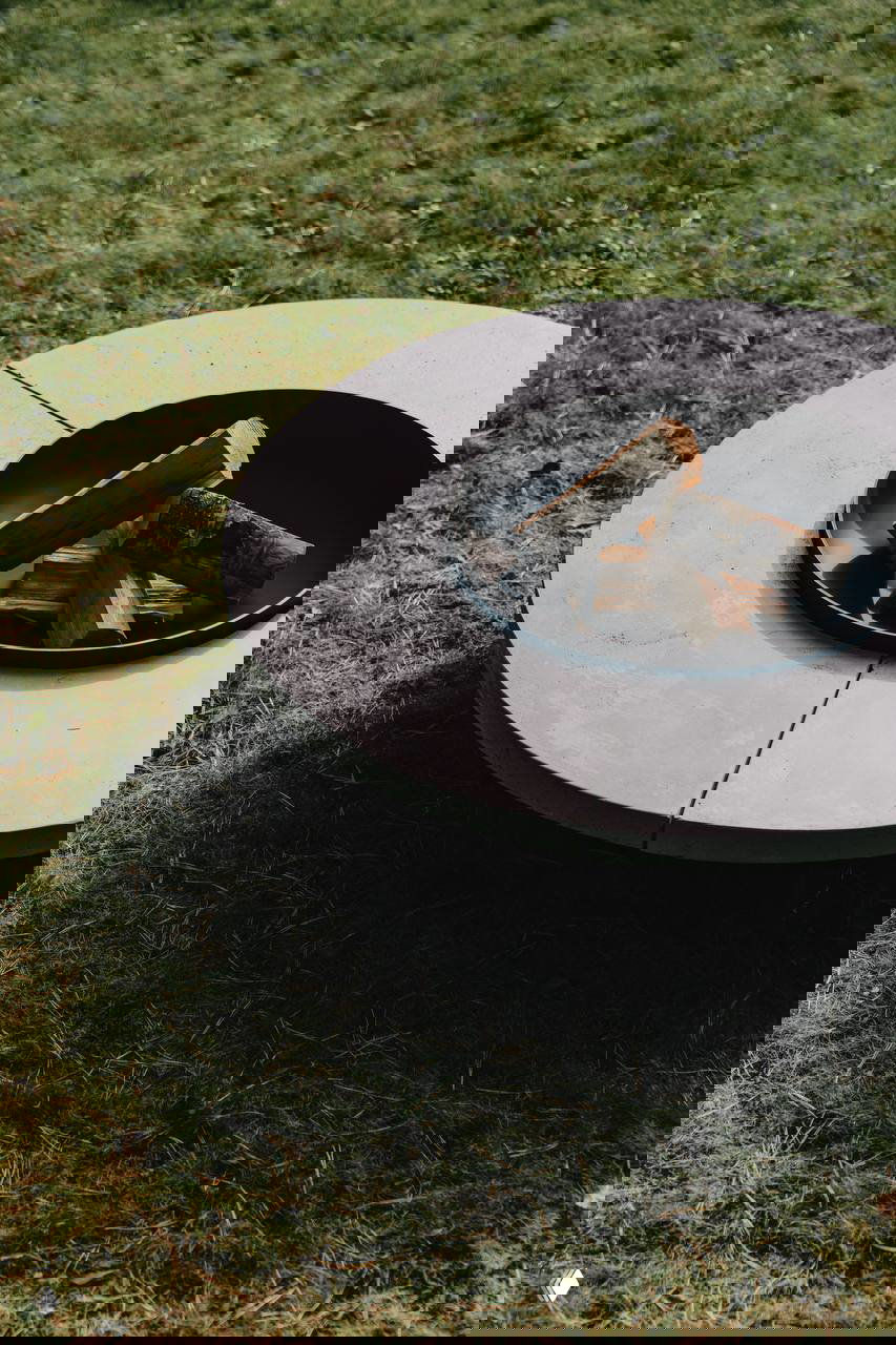 Низкий круглый очаг с бетонной столешницей и металлической чашей, внутри дрова; элемент уличной коллекции BOCA ROOM на траве.