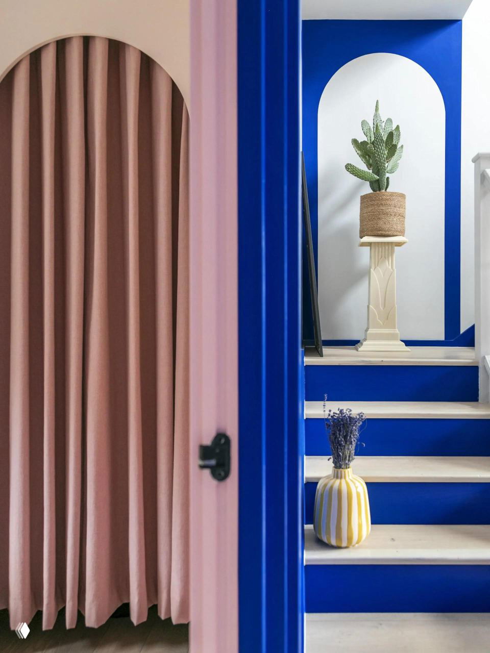 Арочный проём и ступени, выкрашенные в кобальтовый синий, на переднем плане розовые занавеси и высокий декоративный горшок с кактусом на пьедестале.