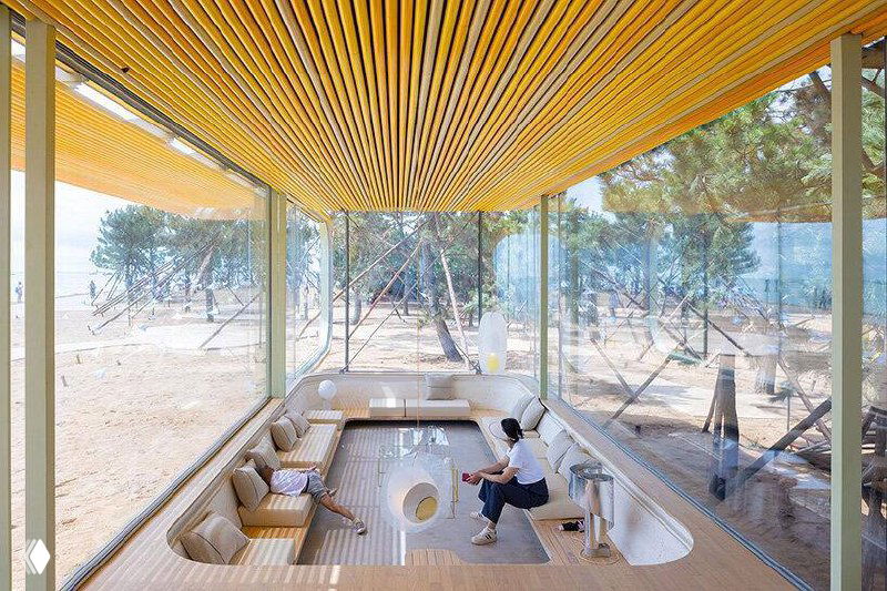 Вид снизу под навесом кафе: решётчатый потолок, прозрачные стены и открытые проходы вдоль пляжа создают лёгкую и светлую атмосферу.