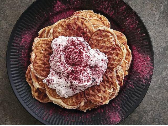 Порция воздушных вафель, украшенных сливочным кремом и ягодным порошком, на тёмной декоративной тарелке.