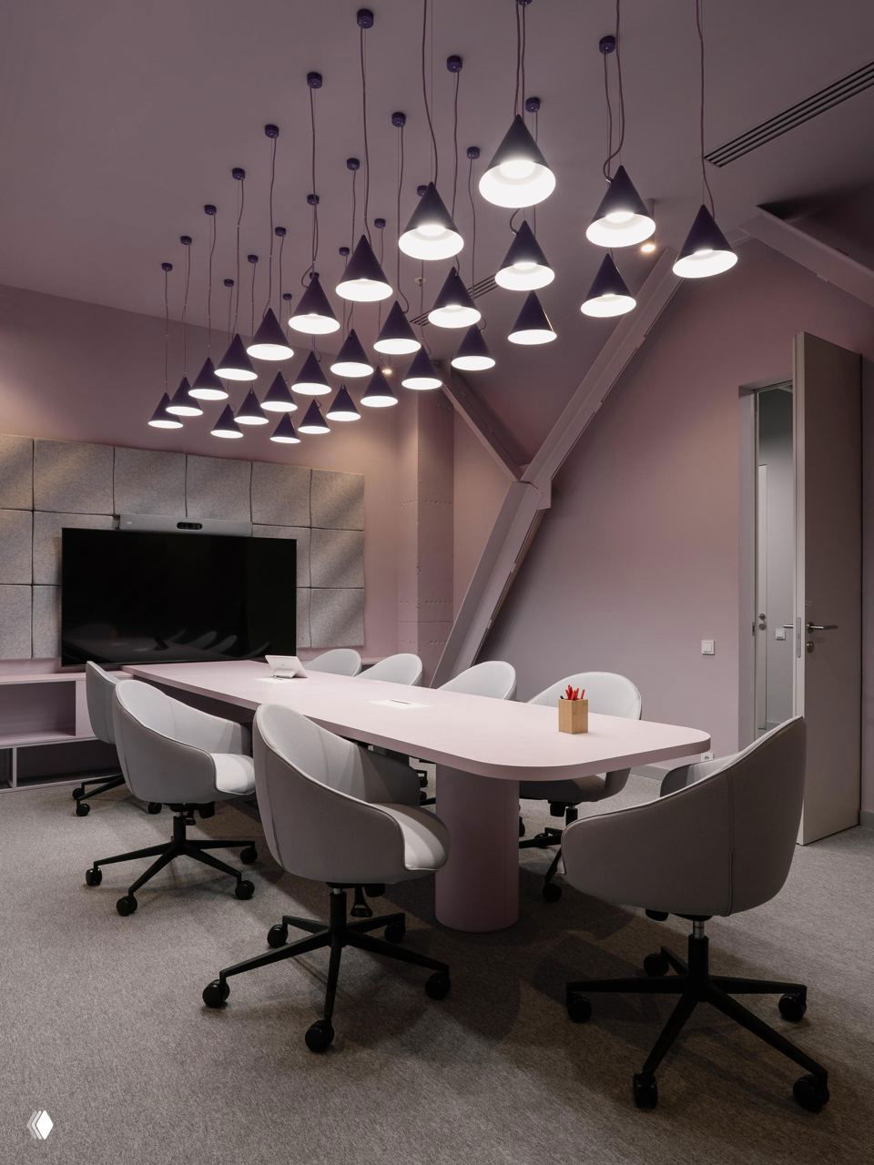 Конференц-зал с длинным столом и мягкими креслами на роликах, нежно-розовые стены и множество подвесных светильников над столом.