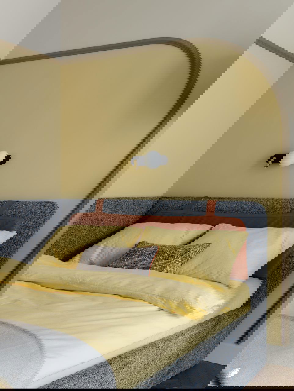 Кровать с жёлтым постельным бельём и текстильным изголовьем с терракотовой вставкой; мягкие формы и настенный светильник в небольшой спальне.