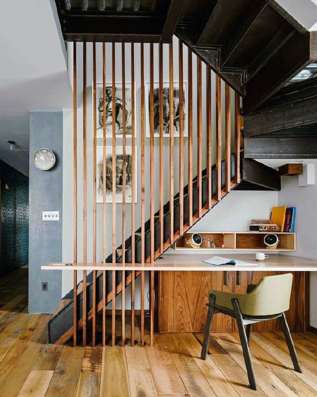 Длинный встроенный стол под лестницей с вертикальными деревянными планками в роли перегородки; тёплый деревянный пол и продуманное освещение рабочего места.