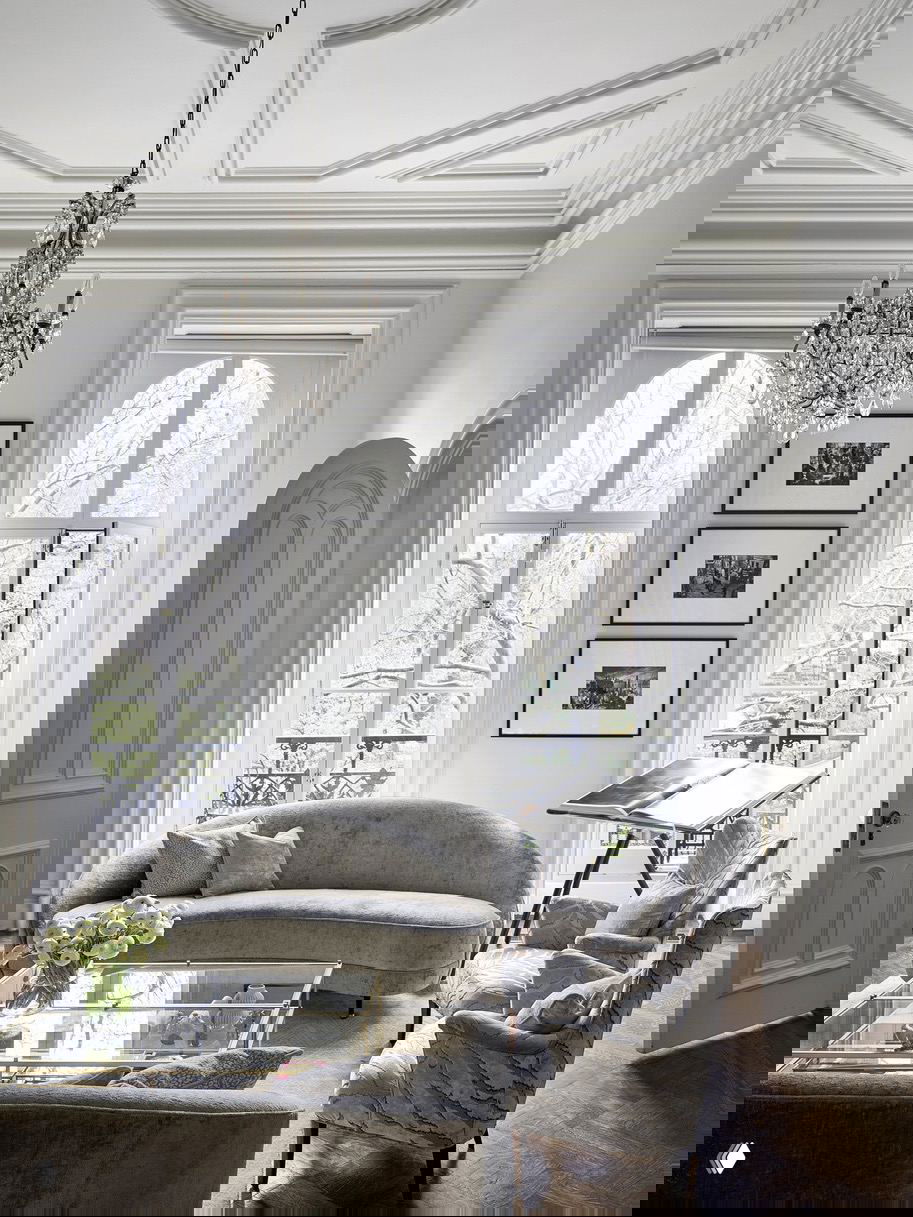 Классическая гостиная с двумя высокими окнами, хрустальной люстрой и серой изогнутой мебелью на паркетном полу у балкона.