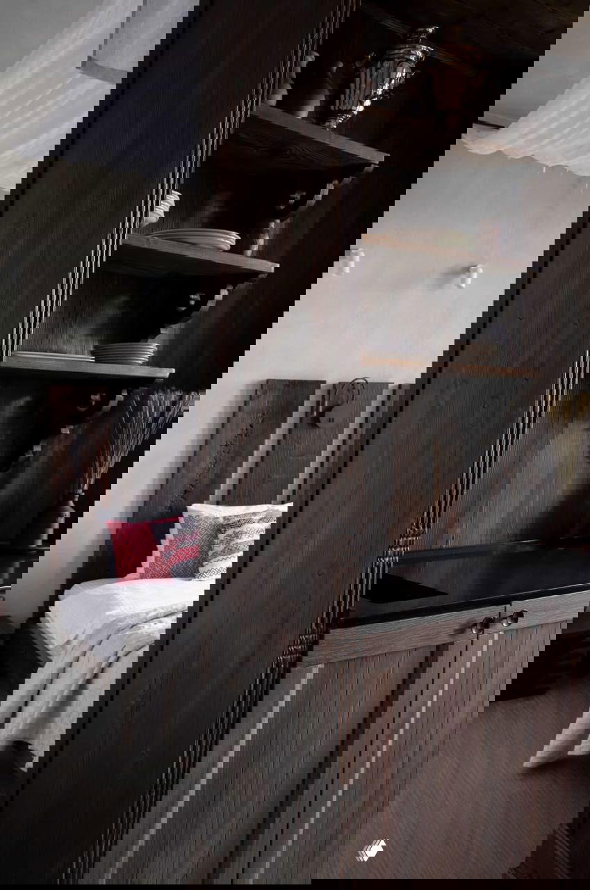 Проход между спальней и кухней с тёмной деревянной мебелью и открытыми полками, текстура дерева и этнические текстильные акценты на кровати.