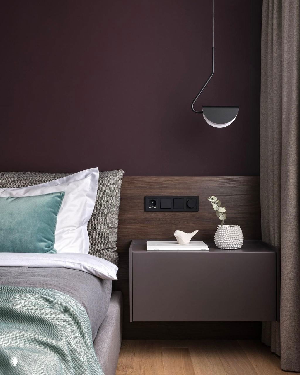 Крупный план прикроватной зоны на фоне тёмно-сливовой стены: современный подвесной светильник, прикроватная тумбочка и предметы декора в минималистичном стиле.
