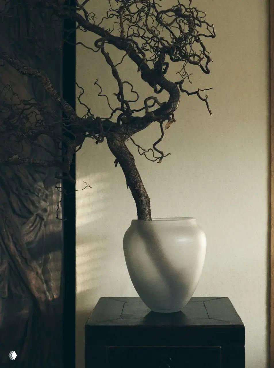 Белая ваза на тёмном постаменте с изогнутой декоративной веткой, кадр в тёплом боковом свете, подчёркивающем фактуру.