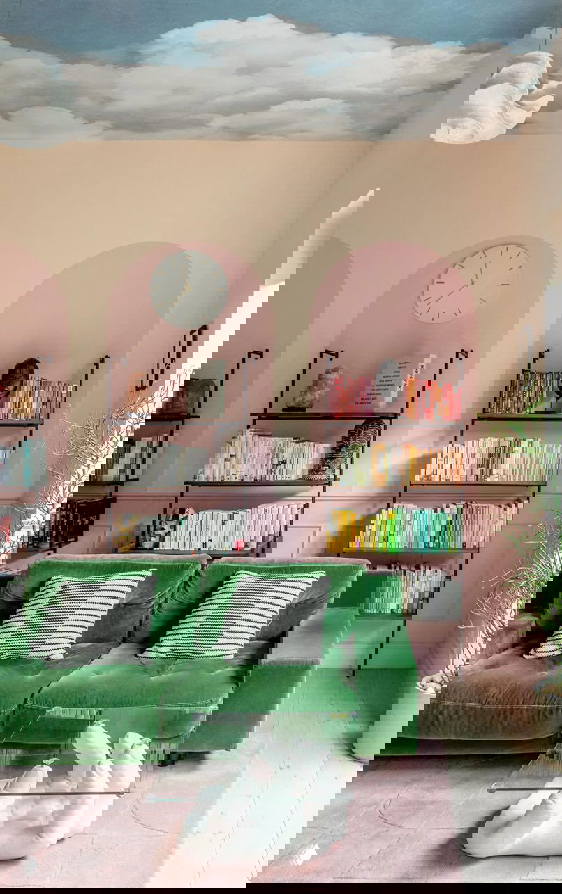 Гостиная с насыщенным зелёным бархатным диваном, розово‑пастельными арками на стенах, книжными стеллажами и стеклянным журнальным столом в виде руки.