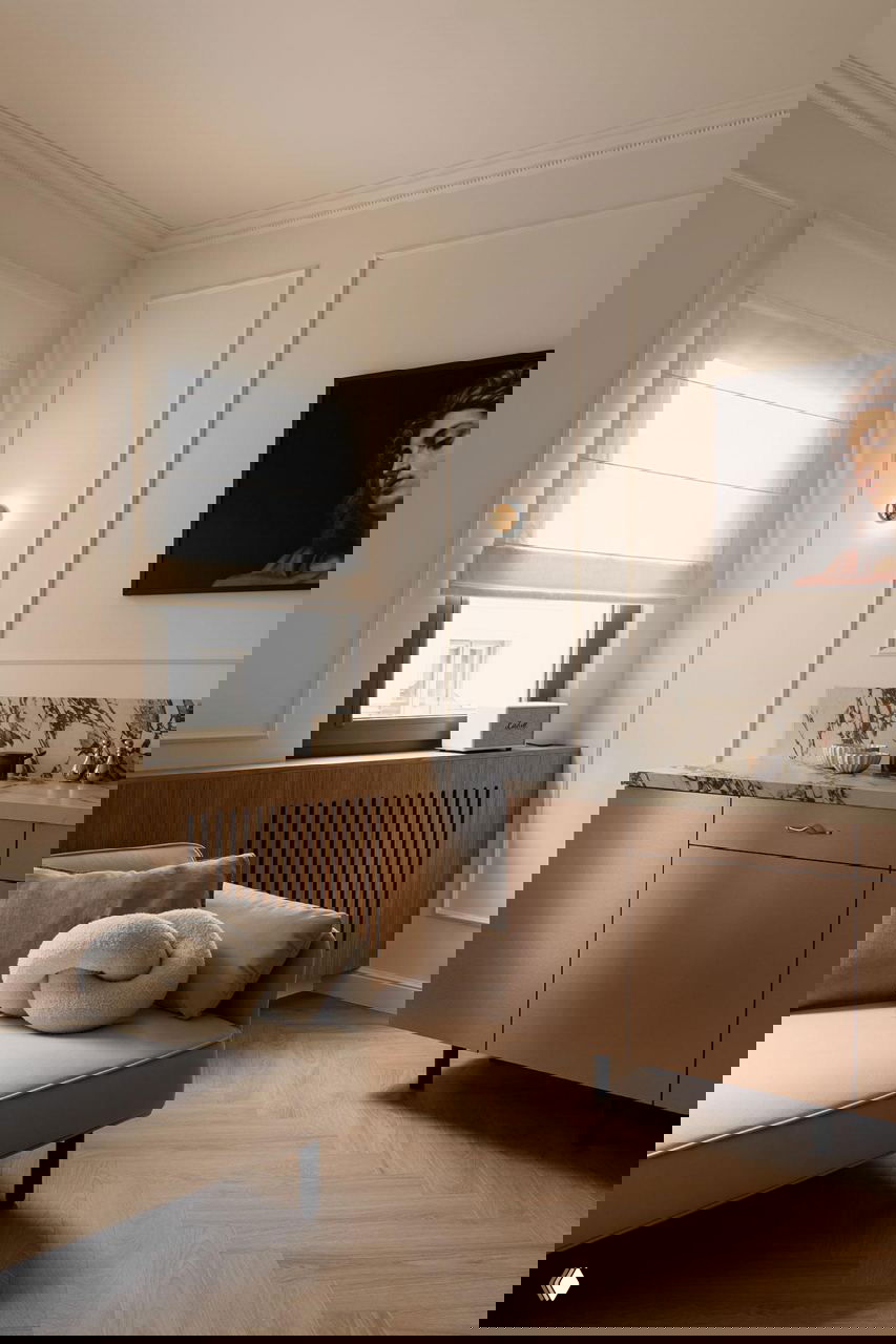 Уютный угол гостиной: высокие окна с рулонными шторами, лепнина на стенах, диван с декоративными подушками и деревянные радиаторные решётки.