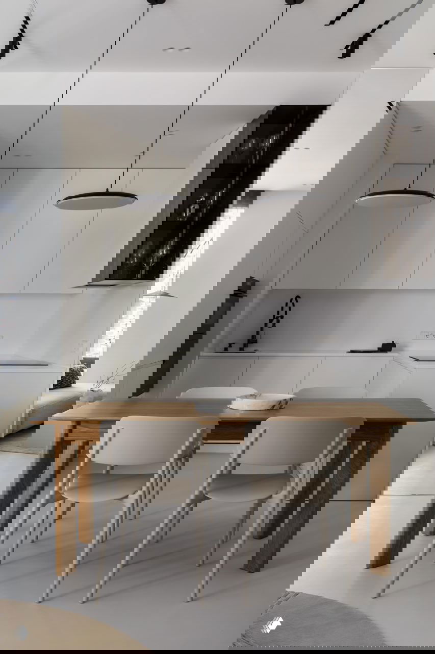 Кухня‑столовая в минималистском стиле: белые встроенные шкафы, ровная рабочая поверхность и деревянный обеденный стол с простыми стульями и подвесными светильниками.