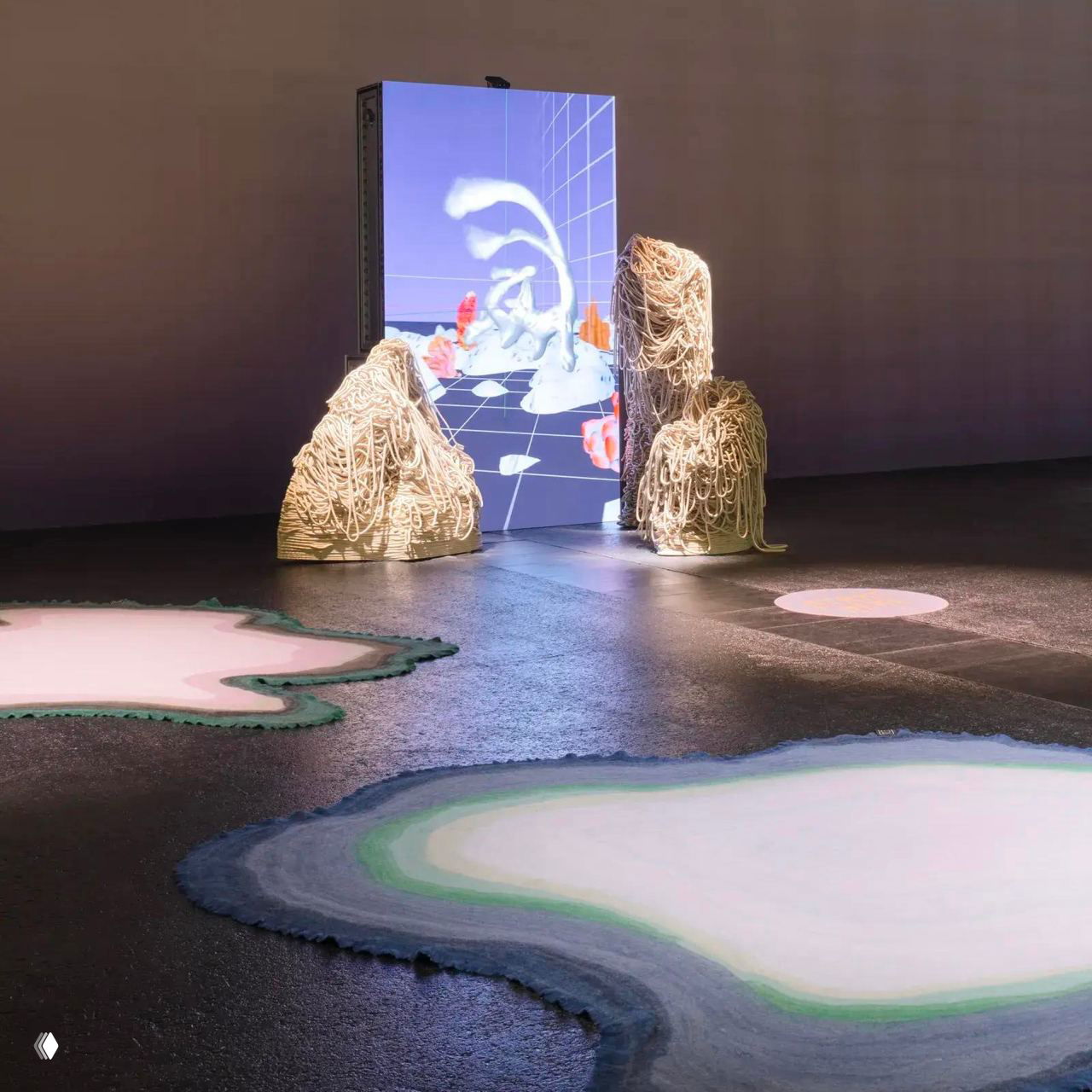 Инсталляция Уркиолы: медиа-экран и текстильные модули, вокруг них органические формы и ковровые пятна из переработанных материалов на полу.