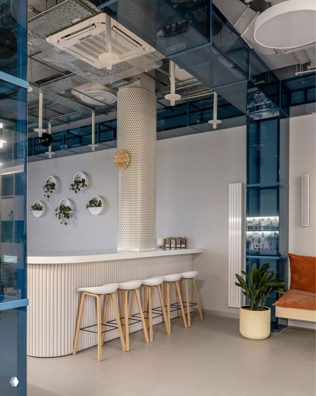 Барная зона офиса с белой гофрированной стойкой, высокими деревянными барными стульями и круглыми кашпо с растениями на колонне.