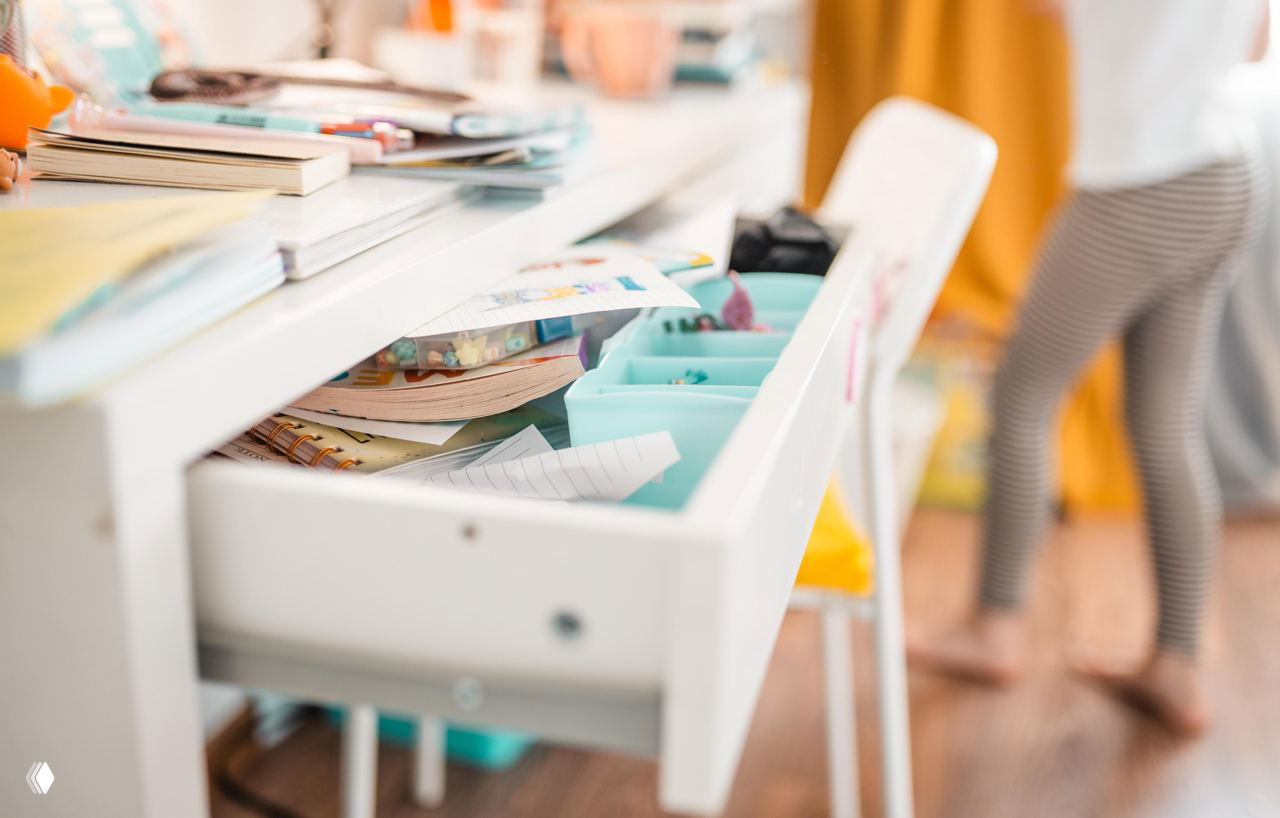 Открытый письменный стол с ящиком, куда сложены тетради, карандаши и органайзеры — пример зоны, требующей ревизии мелочей и упорядочивания.