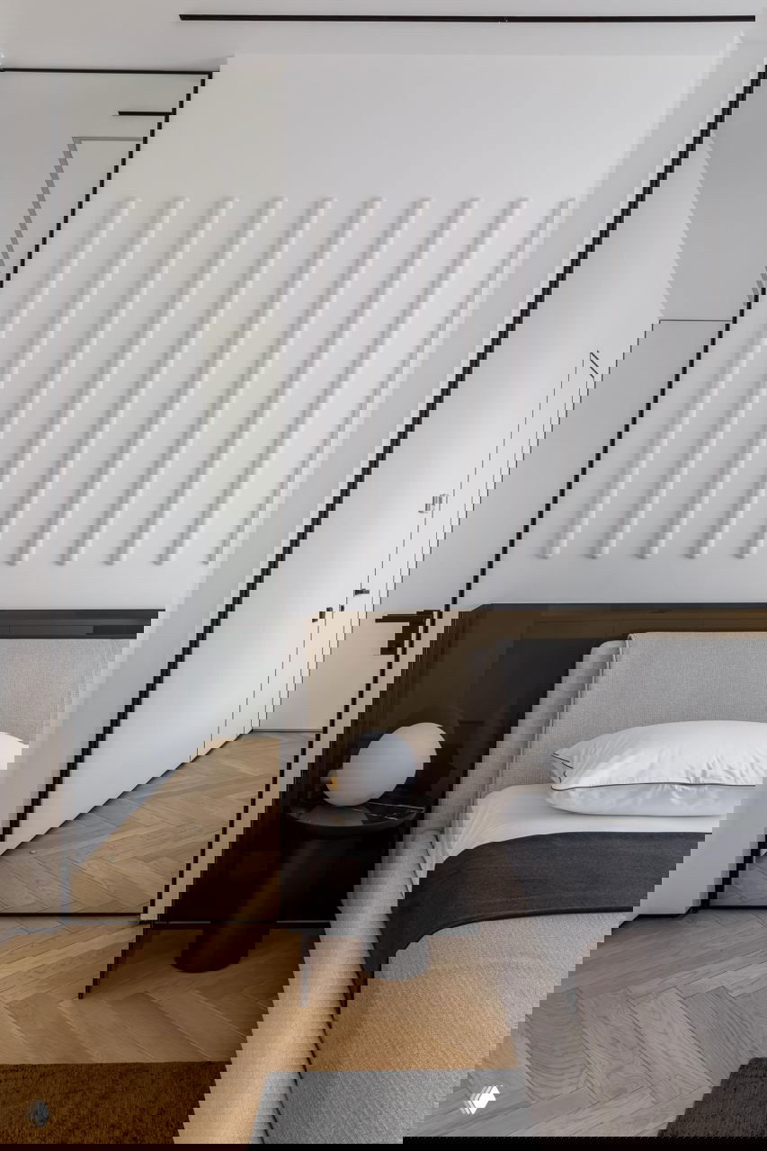 Детали спальной зоны: вертикальные декоративные панели на белой стене, высокое зеркало в раме, низкое мягкое изголовье кровати и паркет «ёлочкой» на полу.