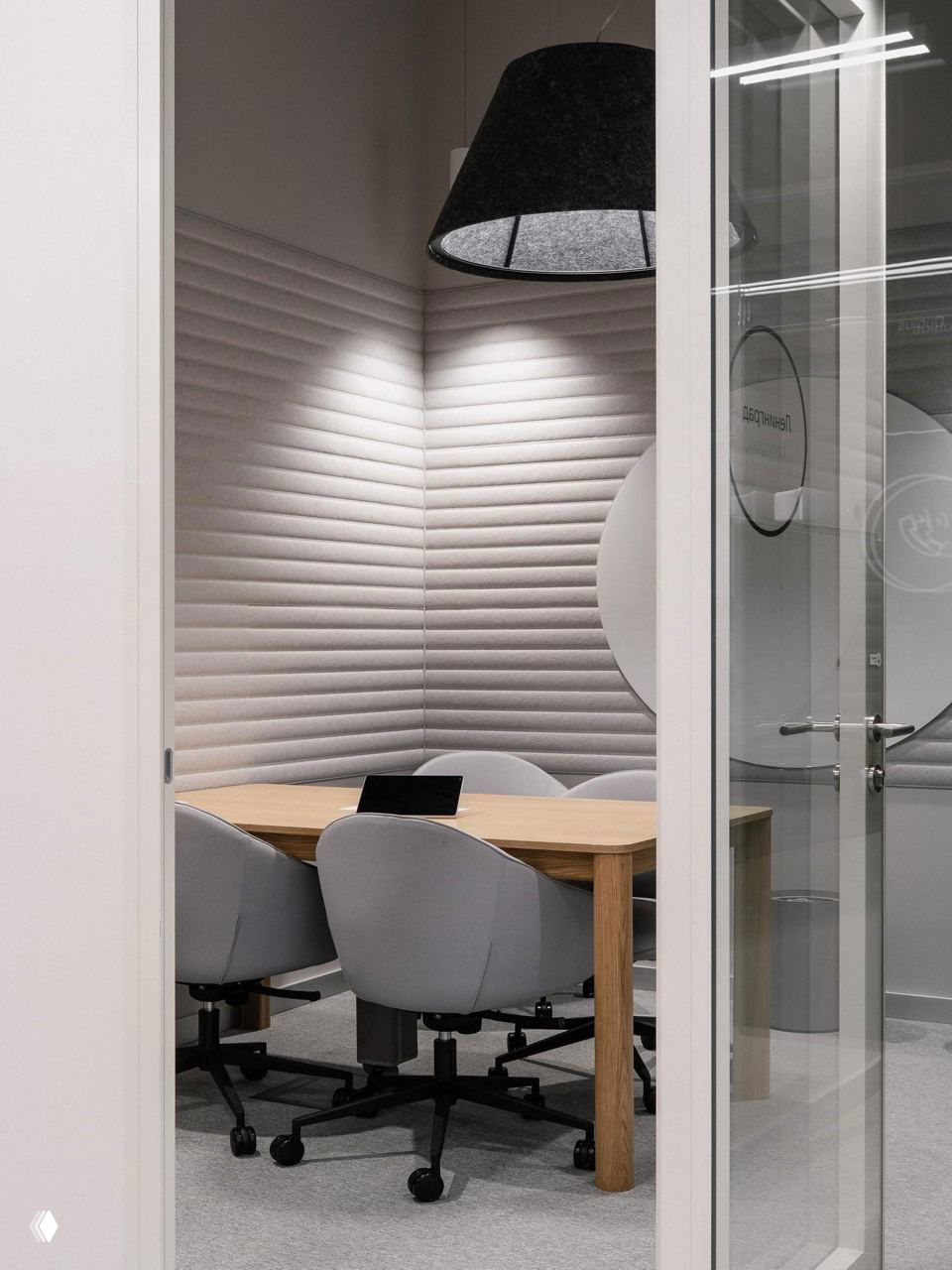 Небольшая переговорная кабинка с акустическими серыми панелями, деревянным столом и четырьмя поворотными креслами на колесиках в офисе.