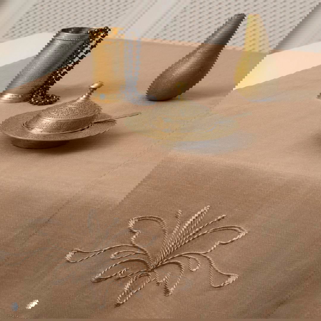 Нейтральная льняная скатерть с тонкой беже‑платиновой вышивкой, на столе металлический подстаканник и груша как декоративный акцент.