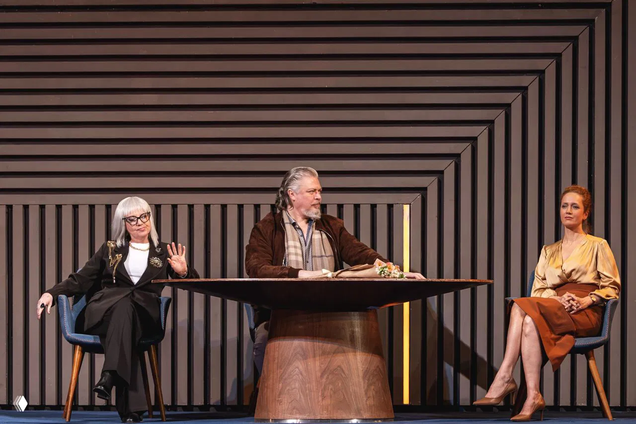 Сцена театрального обсуждения: три актёра вокруг круглого стола на минималистичной декорации, деловое настроение постановки.