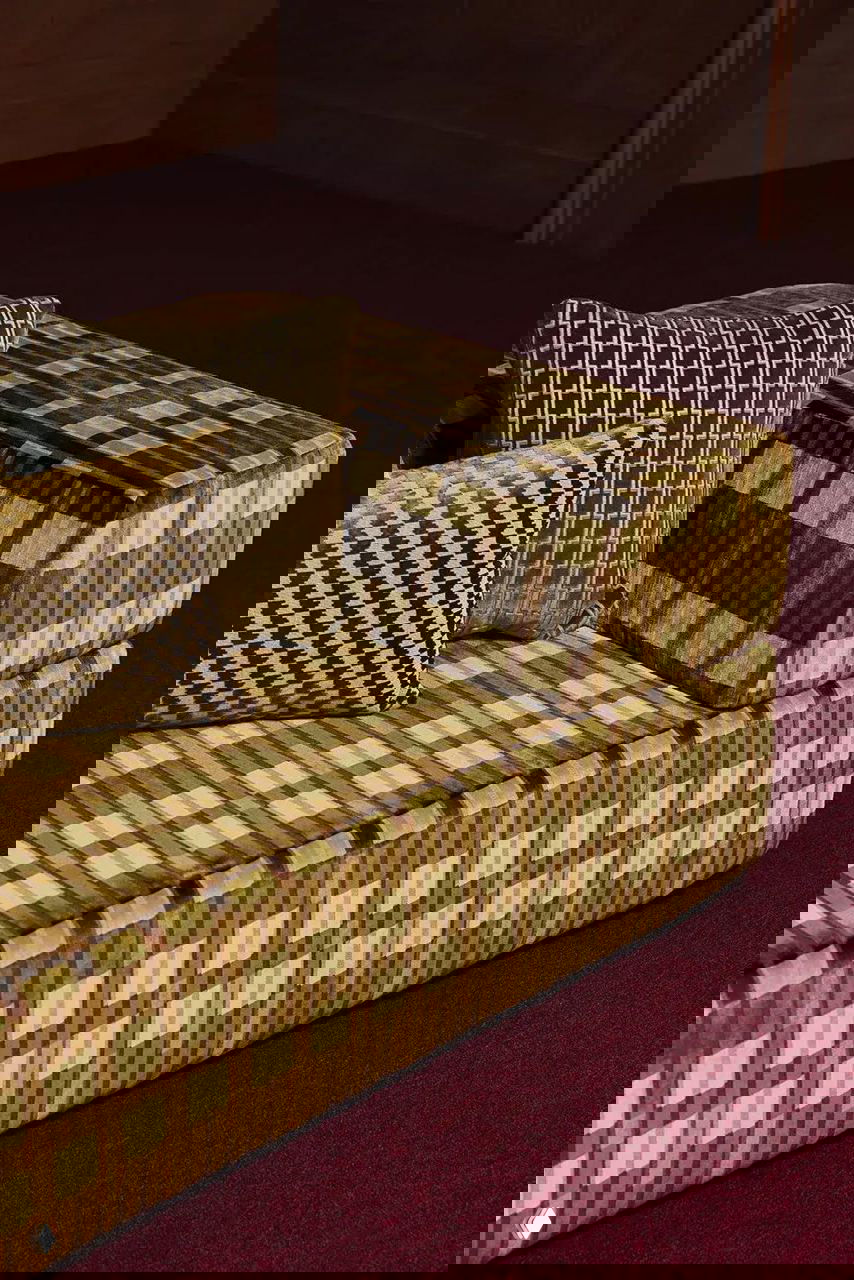 Фрагмент дивана, обитого клетчатой тканью коричневых и бежевых тонов, с декоративными подушками в близком плане на фоне бархатного покрытия пола.