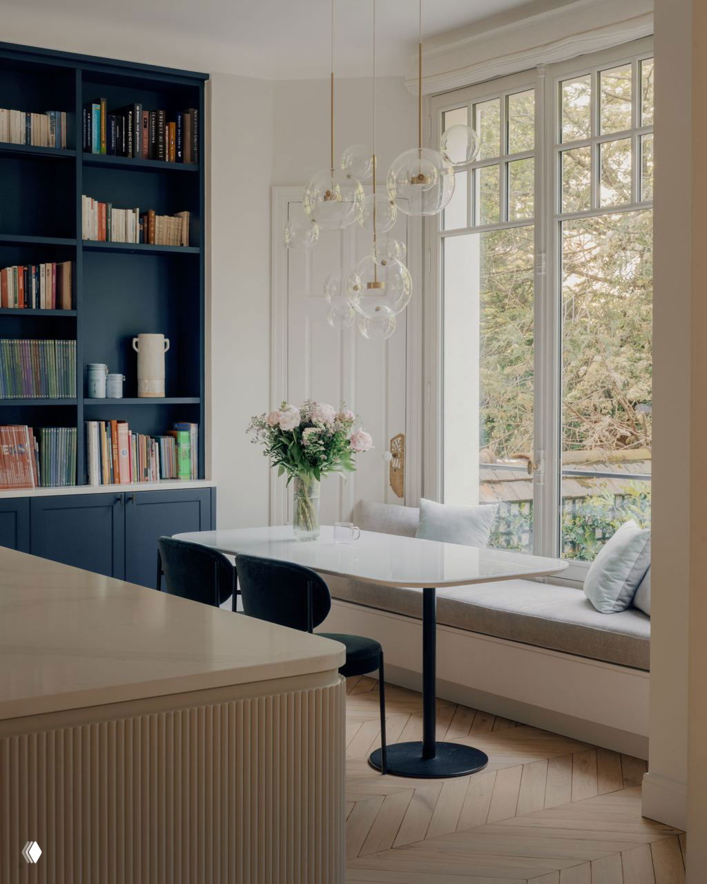 Столовая или уголок для завтрака с большим окном, с синими встроенными шкафами и стеклянной подвесной люстрой над столом.